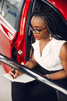 Woman sitting in a red car writing down her budget for car maintenance and auto insurance in a notebook